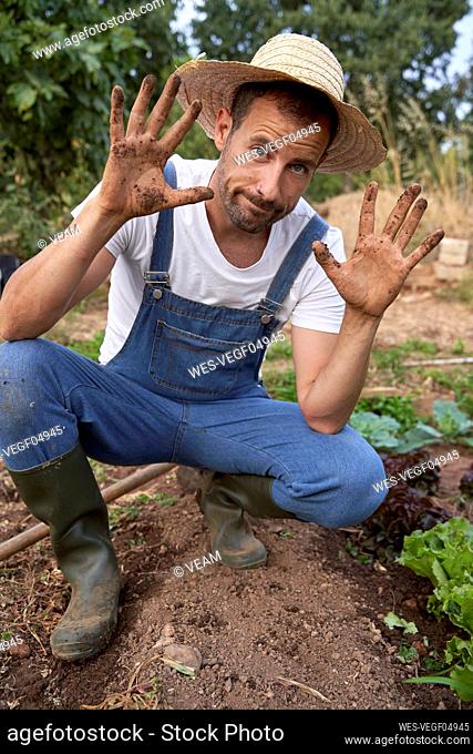 Farm worker in hat showing dirty hands while crouching at agricultural field