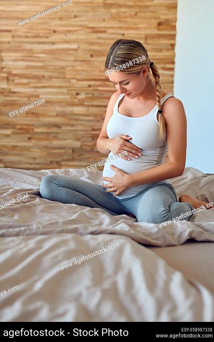 Happy young pregnant woman bonding with her unborn child sitting on a bed cradling her baby bump with her hands looking down with a tender smile of love
