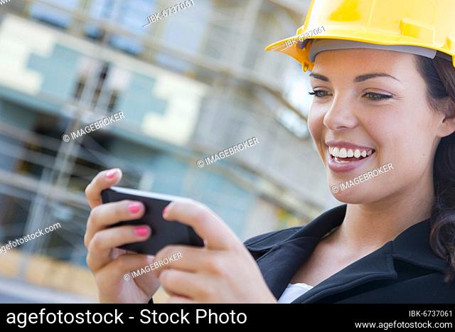 Young professional female contractor wearing hard hat at contruction site texting with cell phone