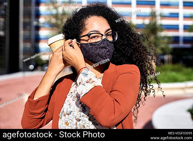 Woman wearing protective face mask while holding reusable bamboo cup in city during sunny day