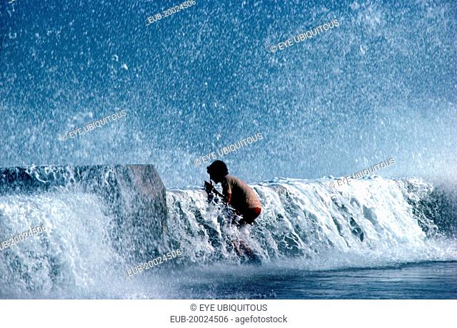 Young boy standing on pavement with waves crashing over the sea wall