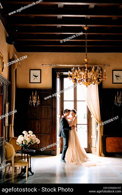 Groom is dancing with bride in the room opposite the table with a bouquet of roses in an old villa. Lake Como. High quality photo