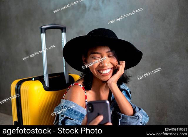 Portrait of smiling young Asian woman with luggage doing selfie photo with her mobile phone isolated over concrete wall. People lifestyle, travel concept
