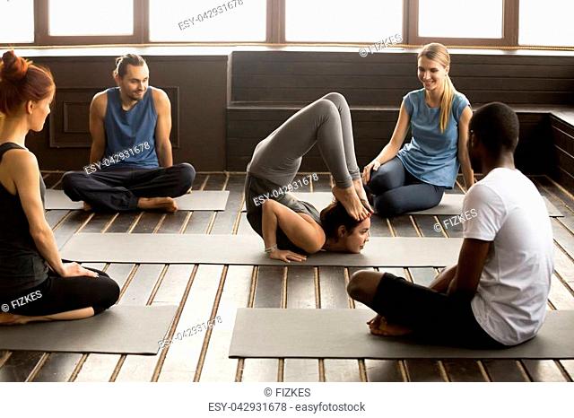 Multiracial diverse people looking at instructor performing acrobatic yoga pose at group training, teacher showing formidable face pose doing ganda bherundasana...