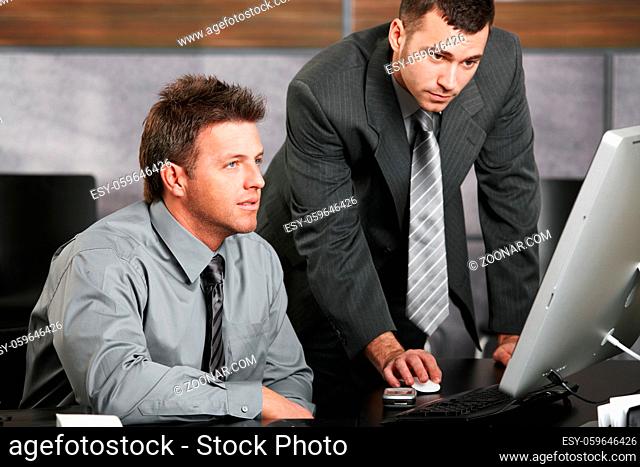 Two businessmen working together with computer at office desk, looking at screen