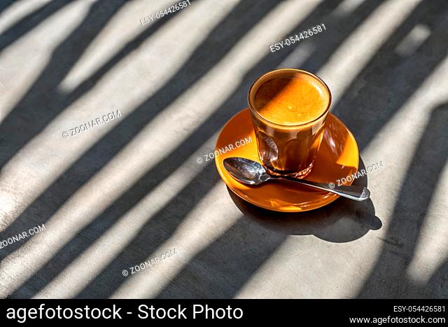 Glass with latte drink on the orange saucer on the table. There is a chrome spoon near it. Sun shines onto them and creates glares and shadow patterns