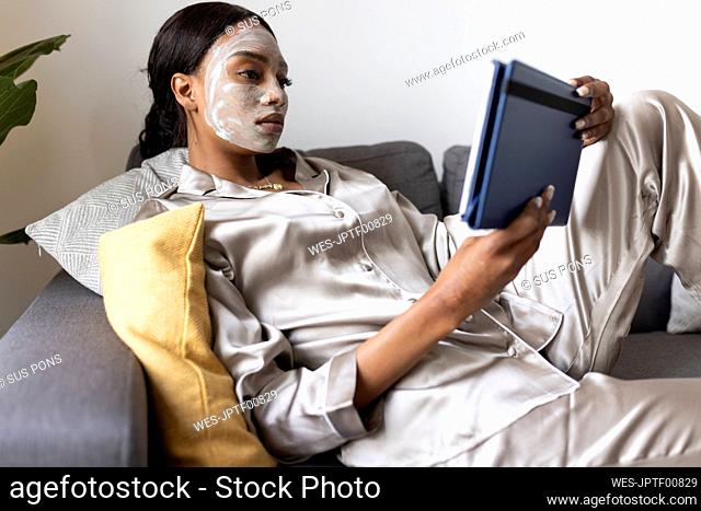 Young woman with beauty mask on face using digital tablet at home