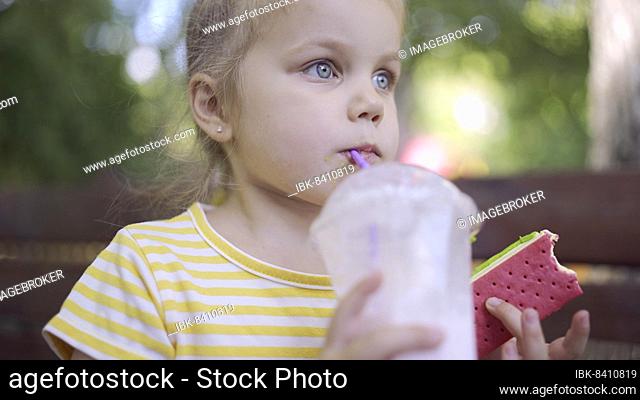 Little girl drinks a milkshake through a straw and smirks at ice cream in her other hand. Close-up portrait of cute child girl sitting on park bench and...