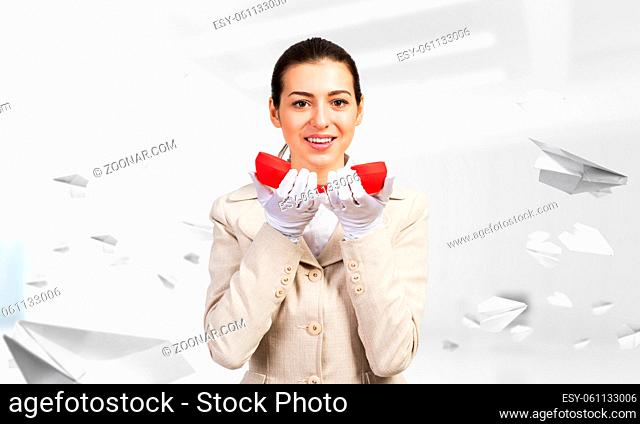 Smiling young woman holding retro red phone in office with flying paper planes. Call center operator in business suit with landline phone
