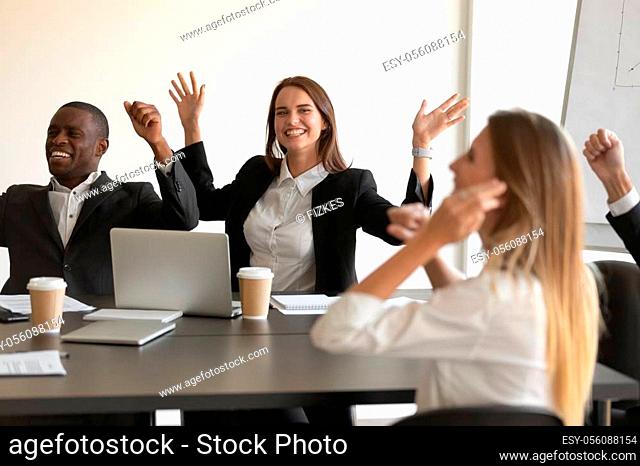 Happy multi-ethnic staff sitting at boardroom desk raising hands screaming with joy feeling excitement, share common success about closing profitable deal