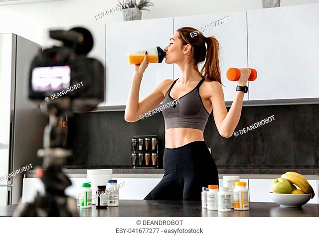 Pretty healthy young girl recording her video blog episode about healthy food additives while standing at the kitchen at home and holding dumbbell