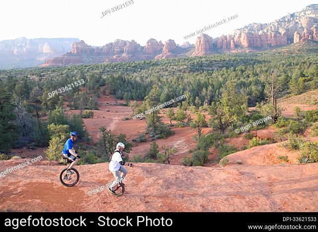 Two men on unicycles ride a rocky ridge in the Arizona desert.; Sedona, Arizona