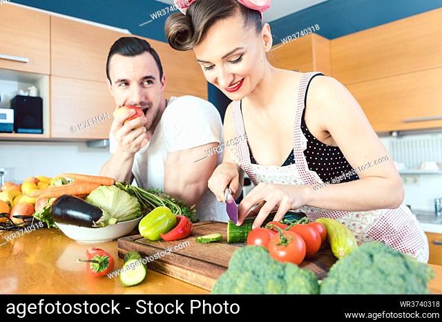 Rather lazy man Is watching his wife preparing the food while eating an apple