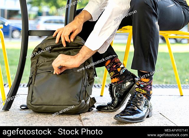Male entrepreneur wearing colorful socks zipping backpack while sitting on chair at sidewalk cafe