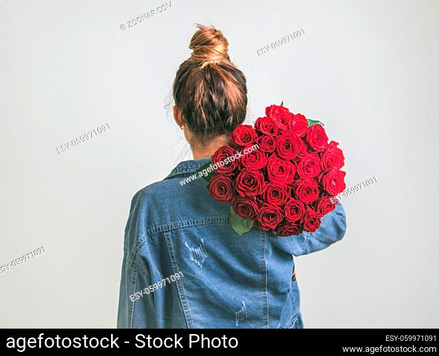 Back view of young woman in denim jacket holding bunch of red roses on shoulder. Girl with bun updo in jeans holding flowers. White background