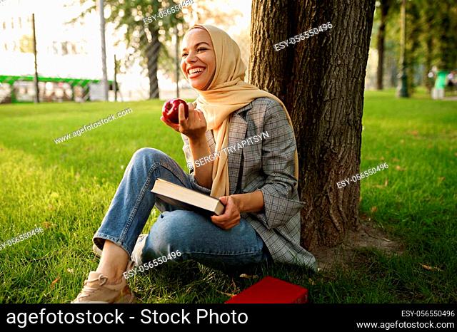 Smiling arab girl in hijab holds apple and textbook in summer park. Muslim woman with books resting on the lawn. Religion and education