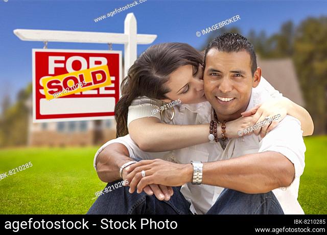 Young happy hispanic young couple in front of their new home and sold for sale real estate sign