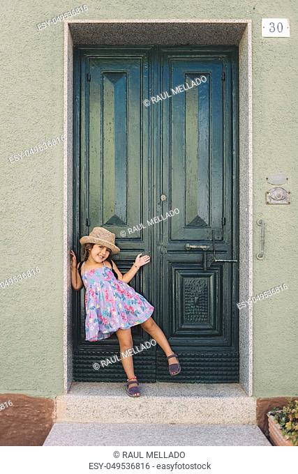 Little girl poses amused and smiling in front of the green door of her house, she wears a lilac summer dress of flowers and a straw hat, vertical image