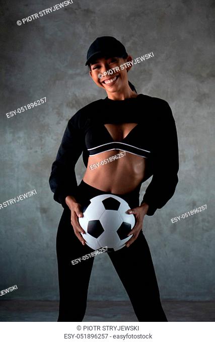 Portrait of beautiful young smiling woman in sports clothes holding soccer ball isolated over concrete background