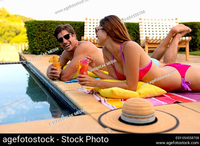 Side view close up of a Caucasian couple wearing beachwear and sunglasses in a garden, lying beside a swimming pool, holding cocktails, sunbathing