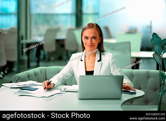 Blond beautiful businesswoman working with documents sitting in front of laptop wearing white official suit. Office worker looking at camera with smile while...