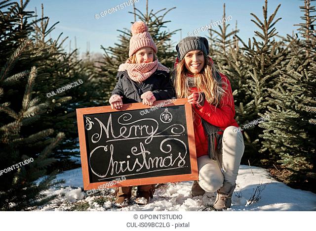 Girl and mother in christmas tree forest with merry christmas sign, portrait