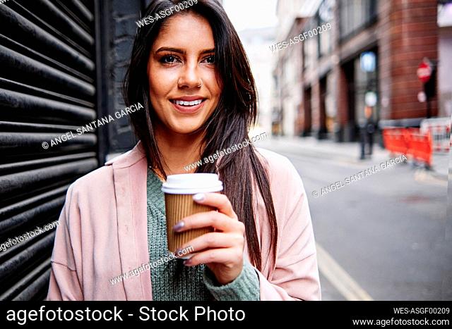Smiling young woman with disposable coffee cup standing by shutter in city
