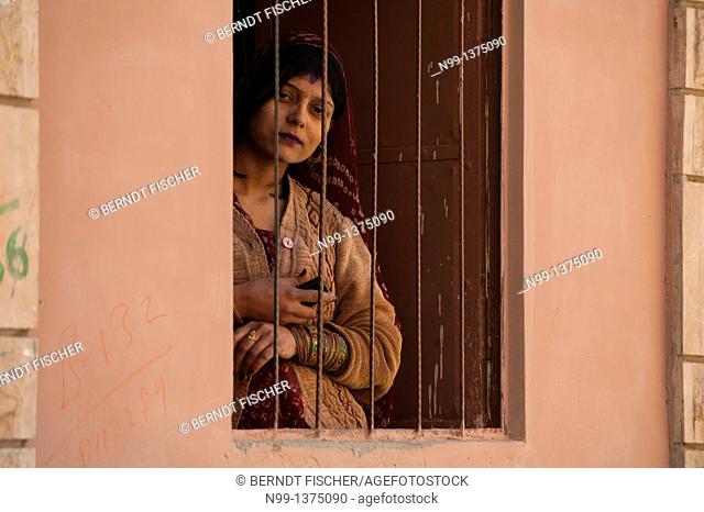 Indian woman with cellphone in her hand, looking through barred window, Phalodi Rajasthan, India