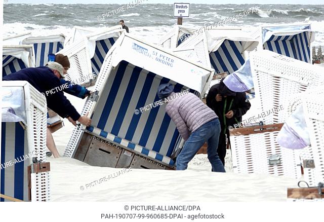 07 July 2019, Schleswig-Holstein, St.Peter-Ording: On the North Sea beach, four people are directing a wicker beach chair blown by the strong wind