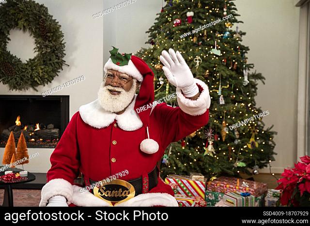 Jolly Black Santa sitting in living room with Christmas tree behind him, waving at camera