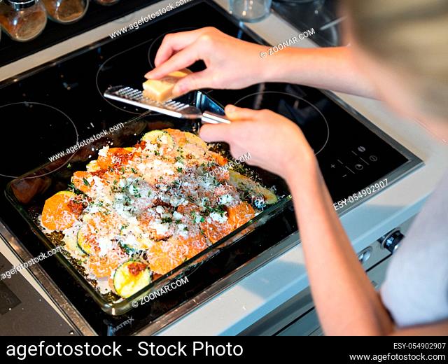 Female chef grinding parmesan cheese on row vegetarian dish ingredients in glass baking try before placing it into oven. Healthy home-cooked everyday vegetarian...