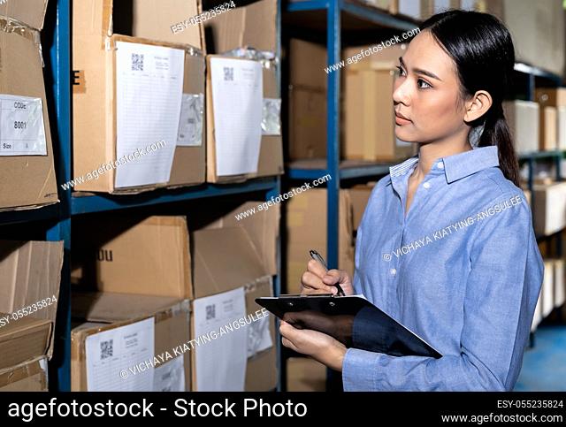 Close up of Asian smart businesswoman working with product inventory and writing on clipboard in distribution warehouse. For business warehouse inventory and...