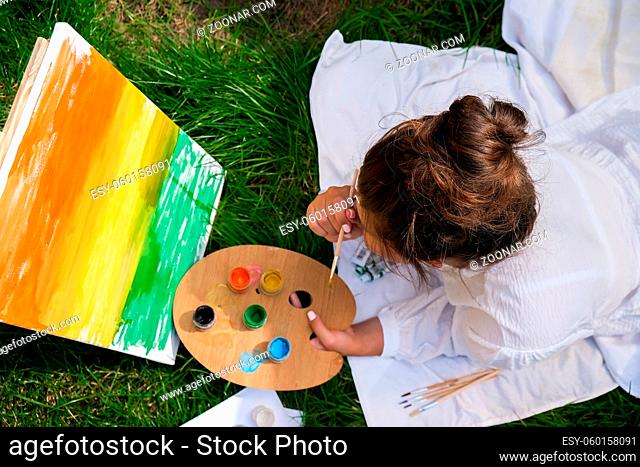 Artist painting on the easel outdoors in the garden. Open air outdoor art workshop. Draw on the canvas with brush and palette sitting on the grass during a...