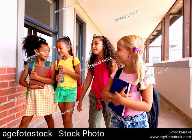 Multiracial elementary schoolgirls with books walking in corridor at school