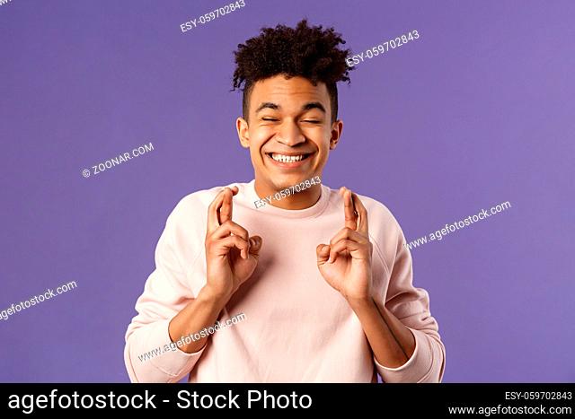 Close-up portrait of happy young hopeful guy anticipating something good happen, cross fingers good luck, close eyes and smiling awaiting miracle
