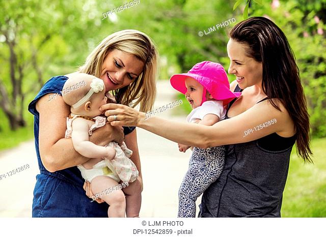 Two young mothers pause while walking in a city park on a warm summer day with their baby girls, who are curious about each other; Edmonton, Alberta, Canada