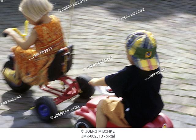 Two children, little boy, little girl, 5-10 years old, playing in the garden, driving a three wheeler and a Bobbycar
