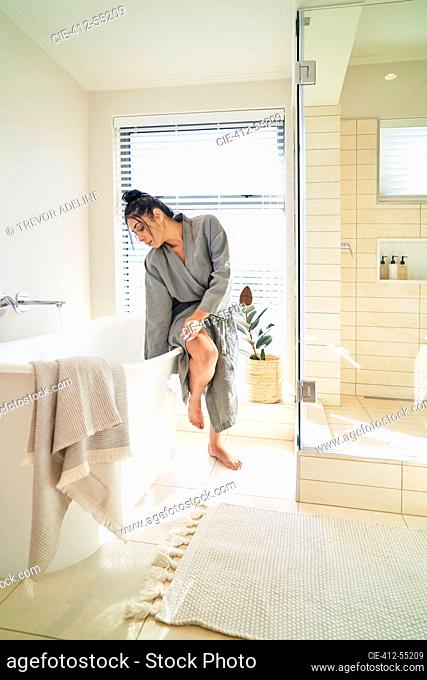 Woman in bathrobe preparing soaking tub for bath in sunny bathroom