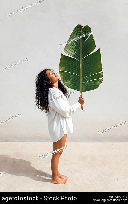 Smiling young woman holding green banana leaf standing by white wall