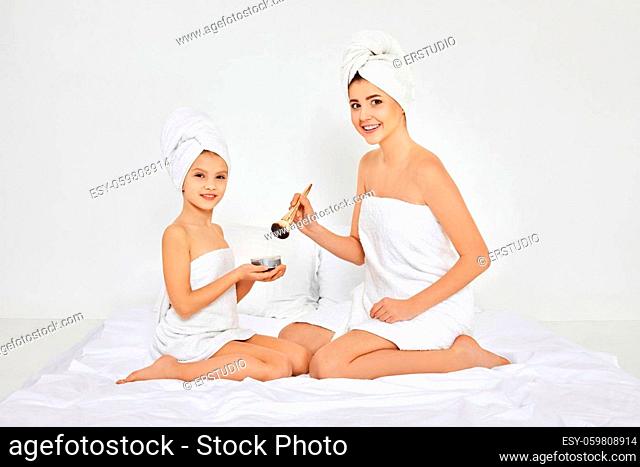 smiling mother and little daughter in white towels doing makeup sitting on the bed in the bedroom