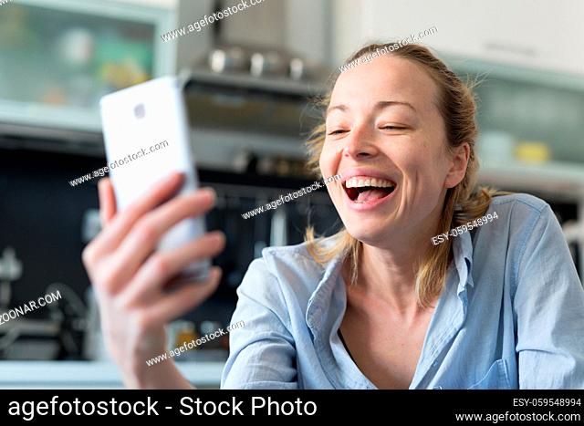 Young smiling cheerful woman indoors at home kitchen using social media apps on phone for video chatting and stying connected with her loved ones