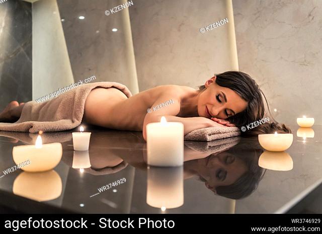 Scented round candle next to young woman on marble massage table during aromatherapy at luxury spa and wellness center