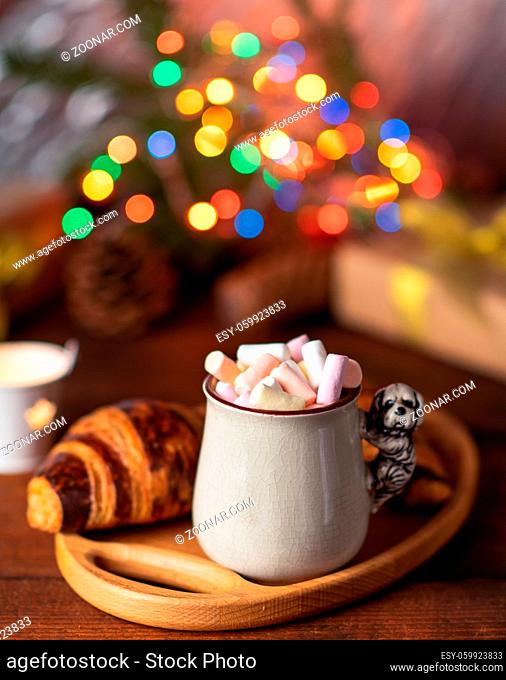 ceramic cup with cocoa and marshmallows and a baked croissant on a brown table, behind Christmas lights