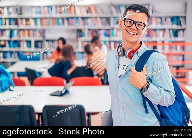 a young student with glasses stands in the school library with a smartphone, headphones and a laptop in his hand