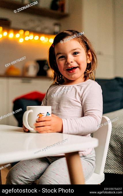 Caucasian toddler girl drinking cocoa from mug at home. Christmas bokeh lights. High quality photo