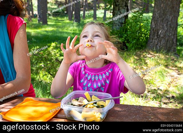 funny four years old blonde girl making fun playing with macaroni, eating pasta from a plastic lunchbox next to her mother in a table picnic in the countryside