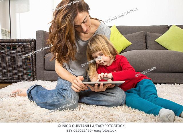 blonde three years old child and mother, with red, green and grey clothes, touching and watching digital tablet to surf internet, sitting on carpet indoor home