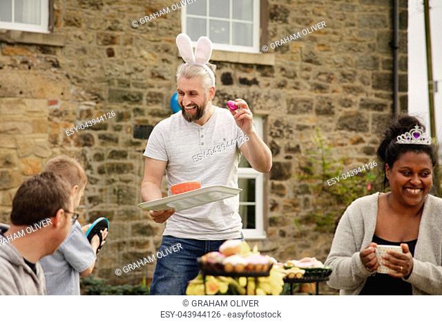 Mid adult man laughing and having fun with other parents at a garden party. He is wearing rabbit ears and holding a chocolate easter egg while smiling