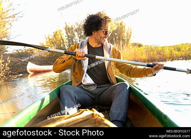 Smiling man wearing sunglasses paddling through oar while sitting in canoe on river