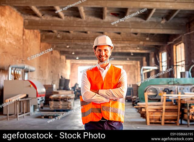 Portrait of young happy and smiling owner of small woodworking factory standing indoors of the shop. Woodworking machinery on the background